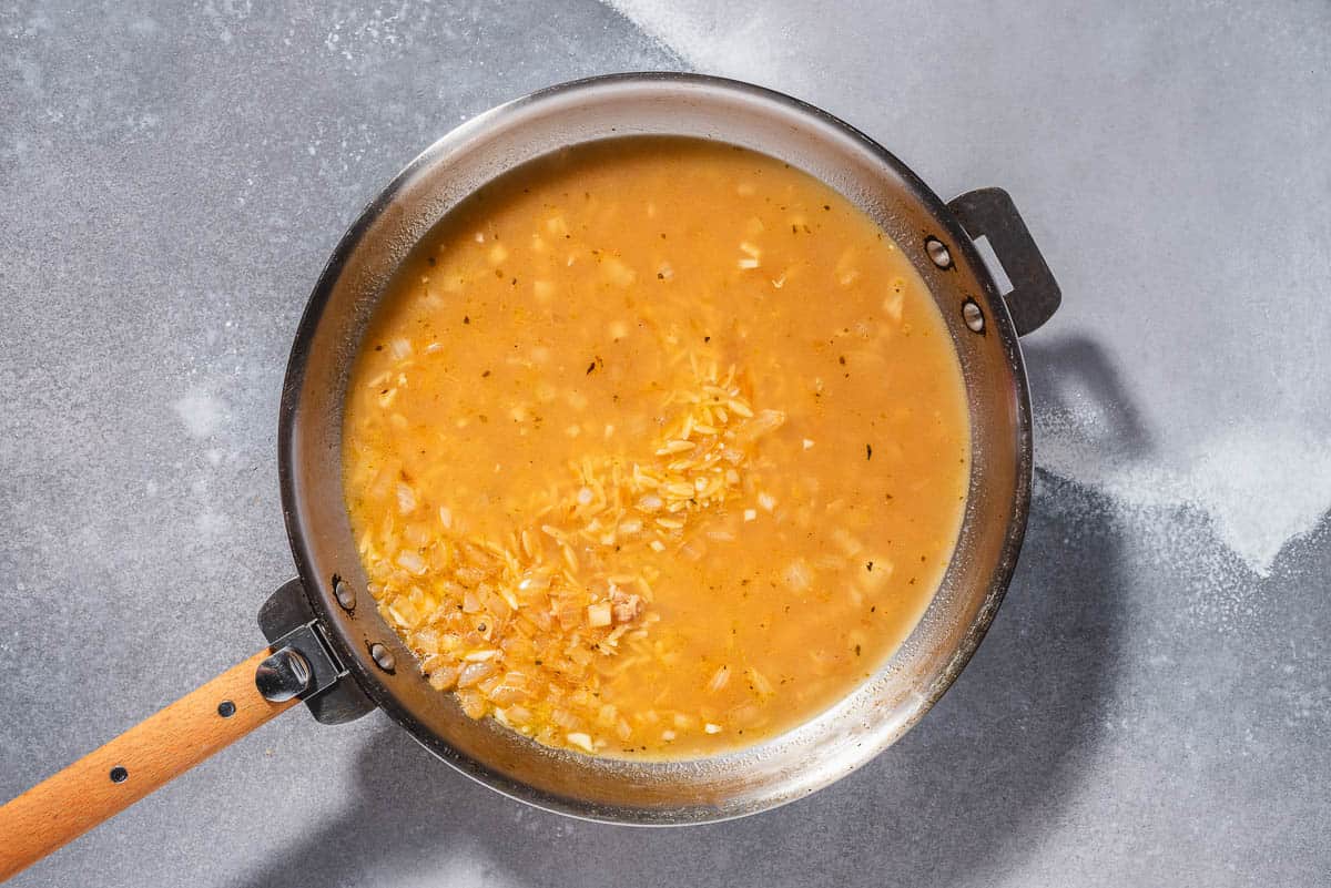 An overhead photo of the orzo and onion in chicken broth in a skillet.