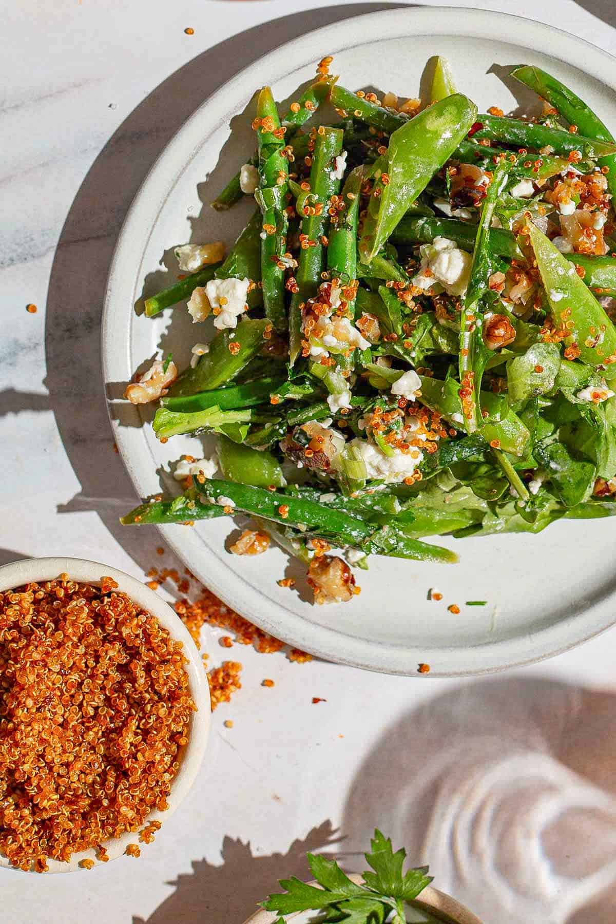 An overhead photo of green beans on a plate garnished with crumbled feta and crispy quinoa. Next to this is a small bowl of the crispy quinoa.