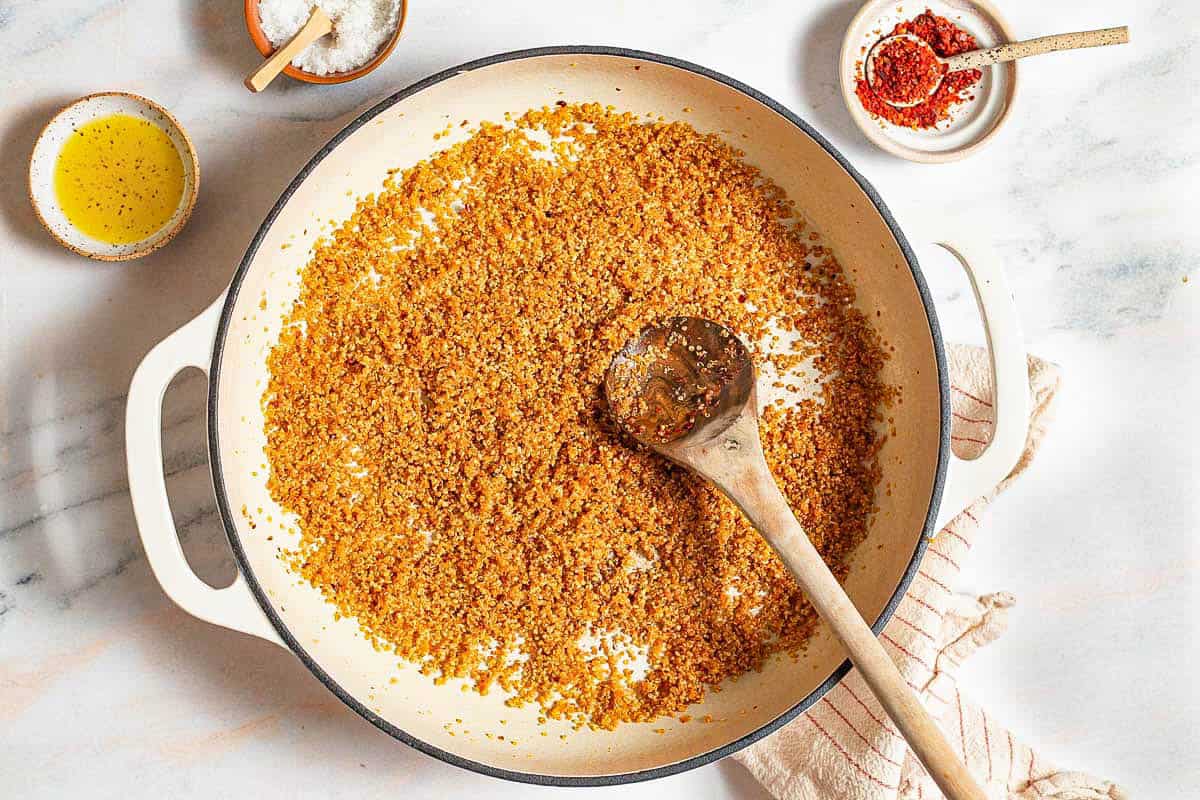 An overhead photo of cooked quinoa being gently toasted in a skillet with a wooden spoon. Next to this is a kitchen towel and bowls of olive oil, kosher salt and aleppo.