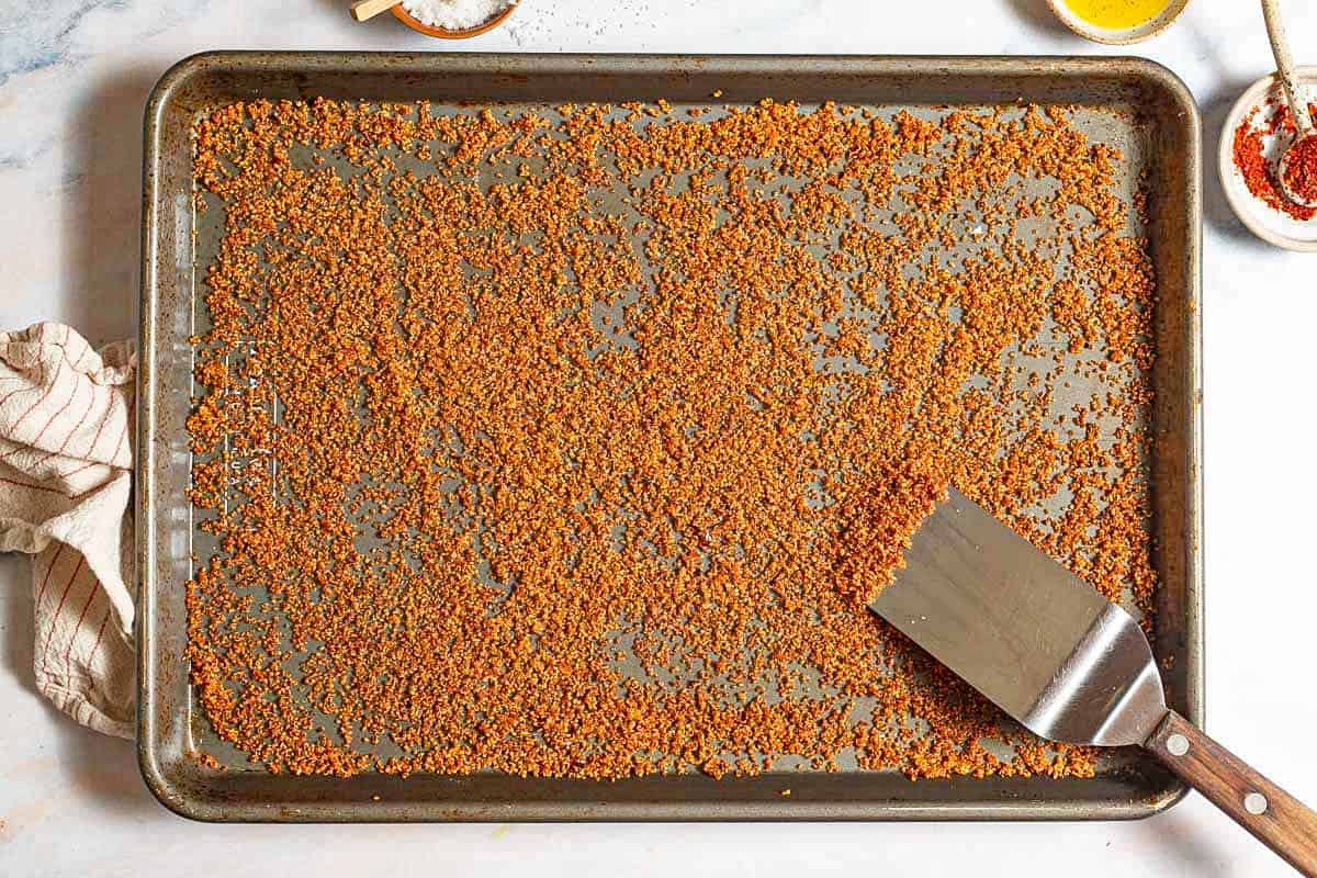 An overhead photo of crispy quinoa spread evenly on a baking sheet with a spatula. Next to this is a kitchen towel and bowls of kosher salt, olive oil and aleppo.