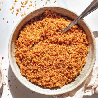 An overhead photo of crispy quinoa in a bowl with a spoon. Next to this is a kitchen towel and bowls of aleppo, olive oil, and kosher salt.