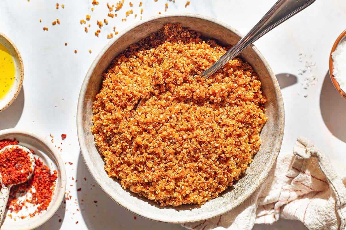 An overhead photo of crispy quinoa in a bowl with a spoon. Next to this is a kitchen towel and bowls of aleppo, olive oil, and kosher salt.