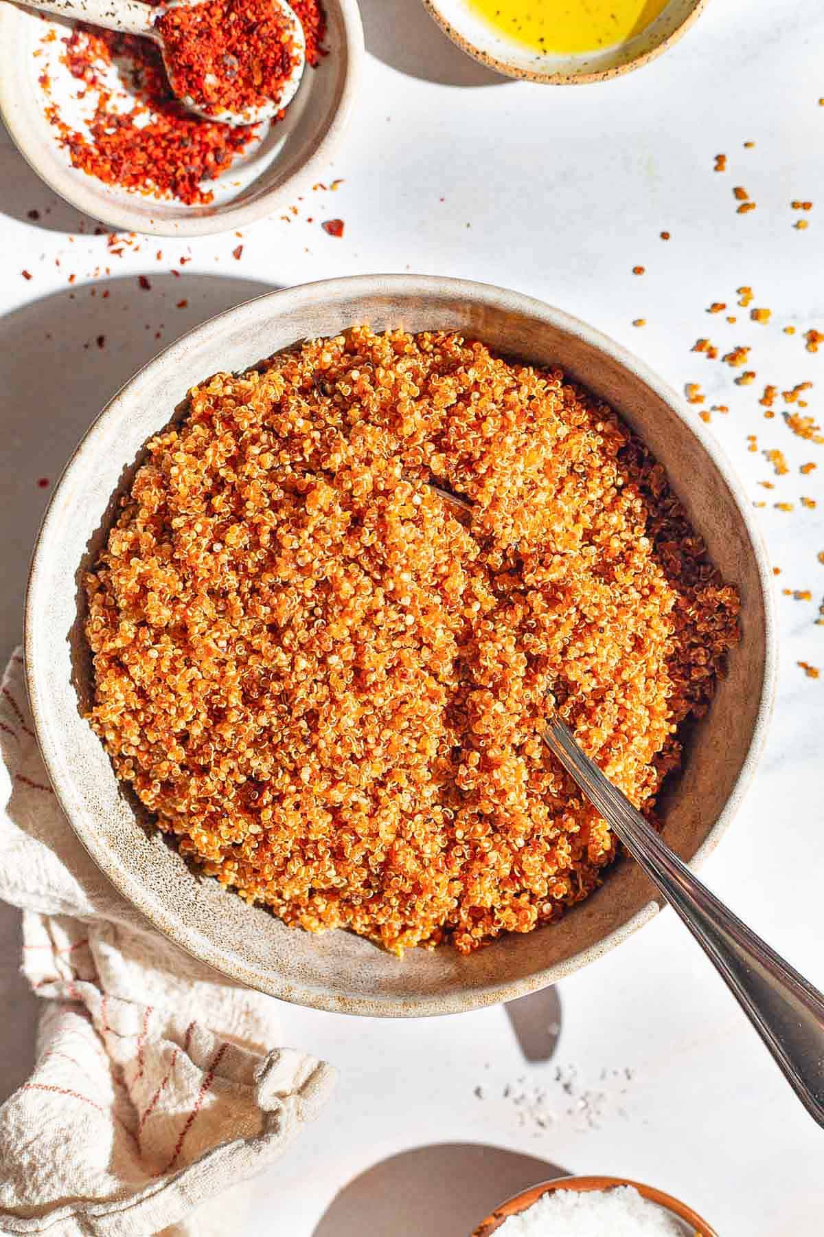 An overhead photo of crispy quinoa in a bowl with a spoon. Next to this is a kitchen towel and bowls of aleppo, olive oil, and kosher salt.