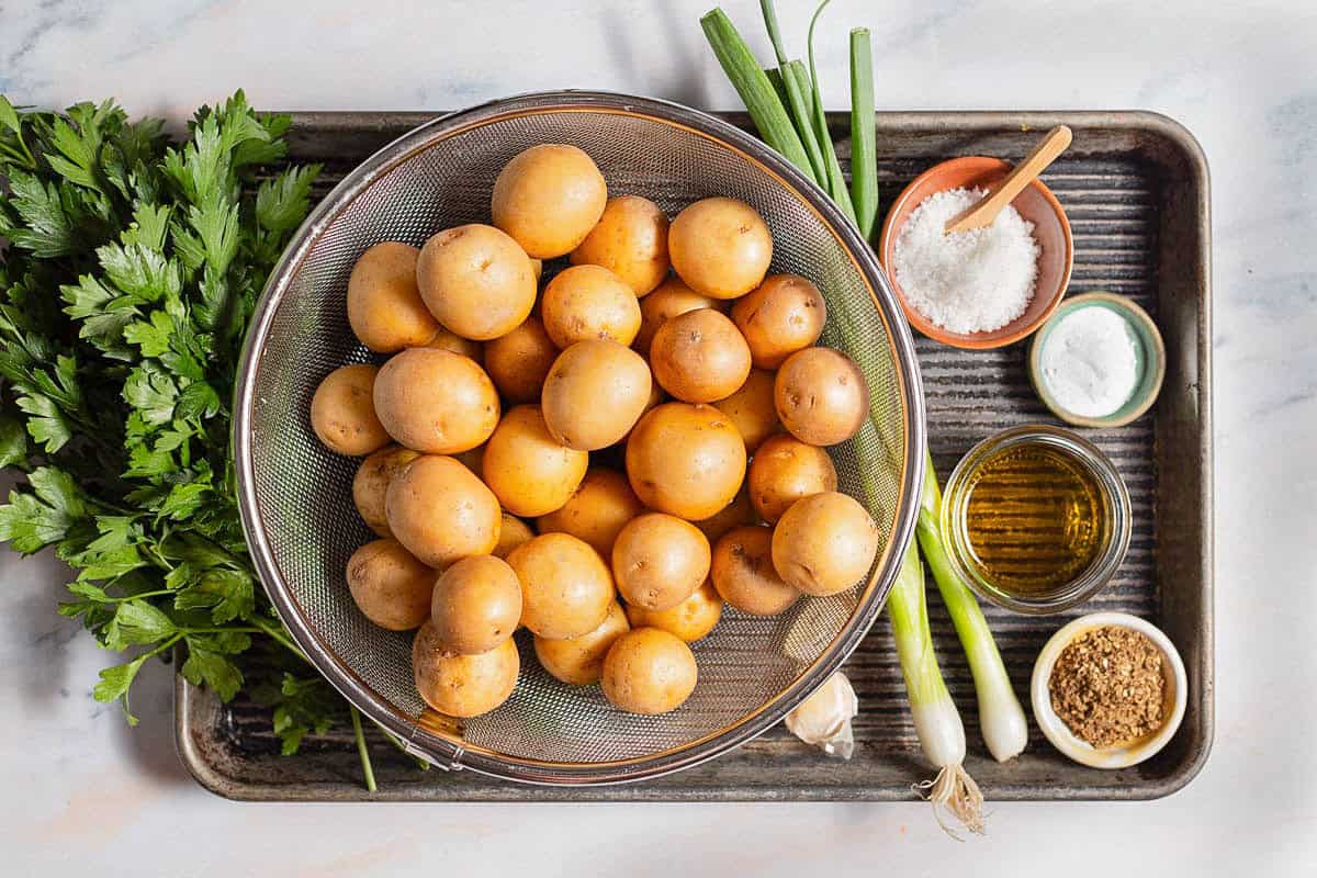 Ingredients for crispy smashed potatoes including baby potatoes olive oil, salt, baking soda, parsley, za'atar, garlic, and green onions.