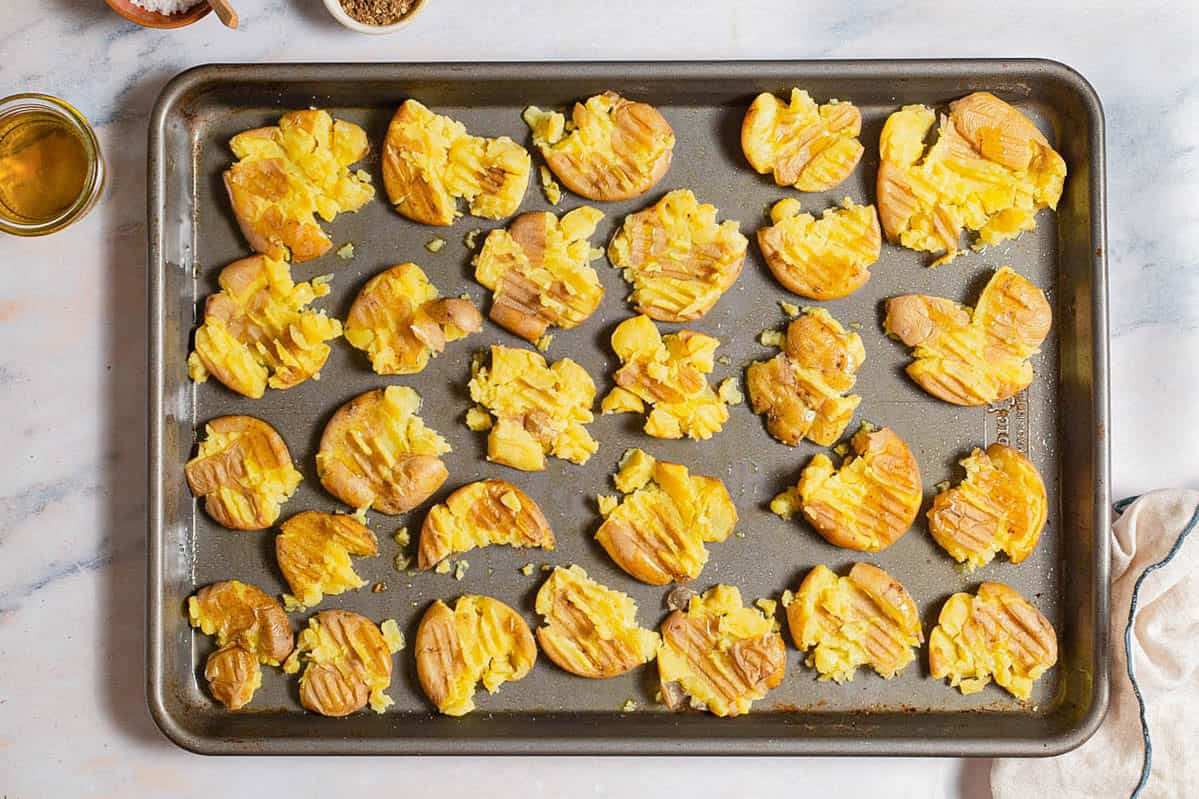 Smashed potatoes spread evenly on a baking sheet. Next to this is a kitchen towel and a bowl of olive oil.