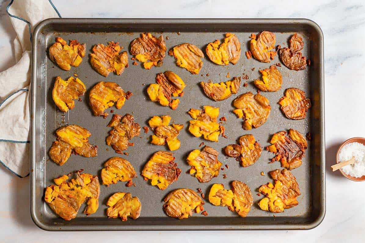 Baked smashed potatoes spread evenly on a baking sheet. Next to this is a kitchen towel and a bowl of salt.