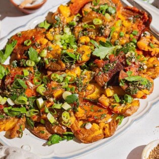 A close up of crispy smashed potatoes on a serving platter with a fork. Next to this are bowls of chopped green onions, kosher salt, za'atar, and parsley.