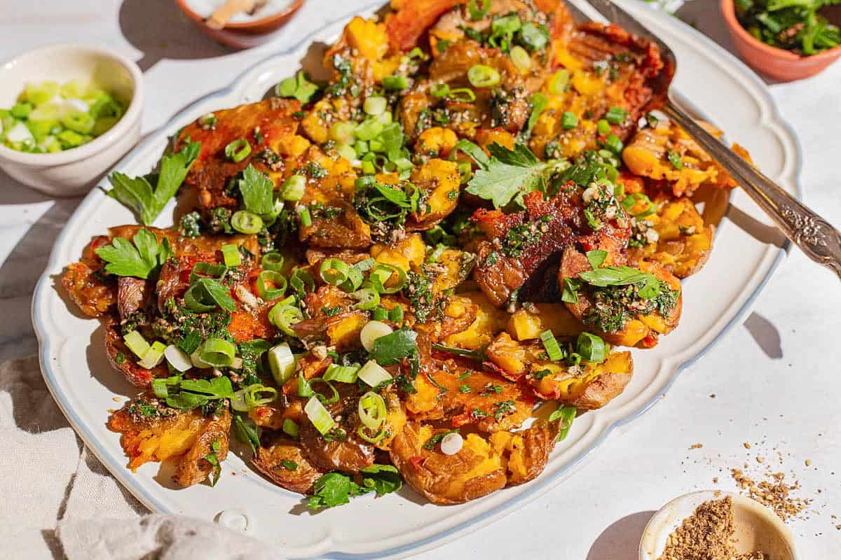 A close up of crispy smashed potatoes on a serving platter with a fork. Next to this are bowls of chopped green onions, kosher salt, za'atar, and parsley.