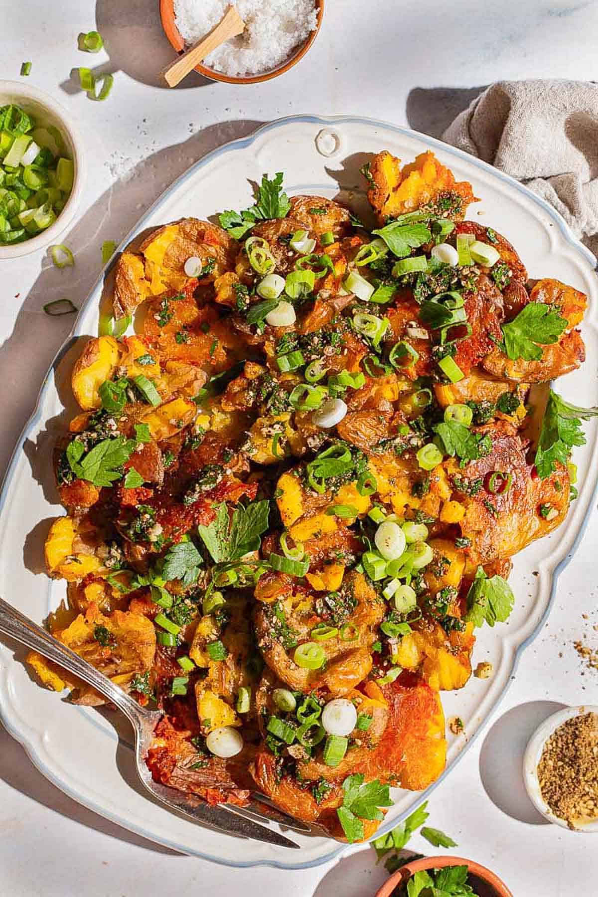 An overhead photo of crispy smashed potatoes on a serving platter with a fork. Next to this are bowls of chopped green onions, kosher salt, za'atar, and parsley.