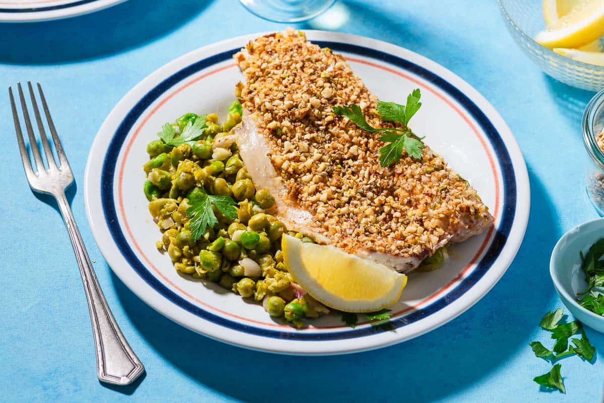 A close up of a serving of baked white fish topped with dukkah on a bed of smashed peas on a plate with a lemon wedge. Next to this is a fork.