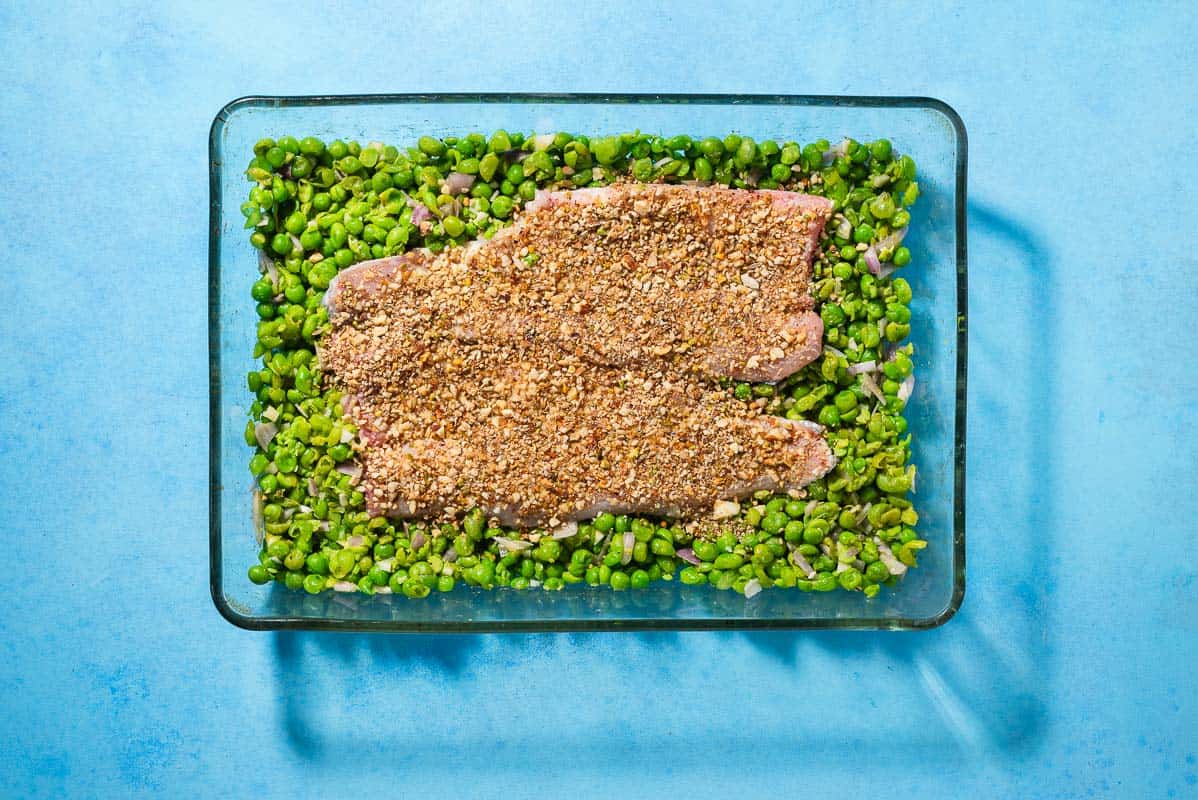 An overhead photo of unbaked white fish topped with dukkah on a bed of smashed peas in a baking dish.