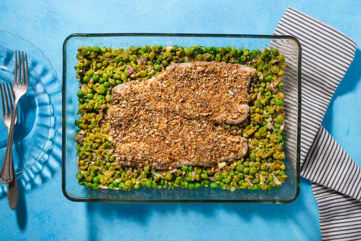 An overhead photo of baked white fish topped with dukkah on a bed of smashed peas in a baking dish. Next to this is a kitchen towel and a stack of 2 plates with forks.