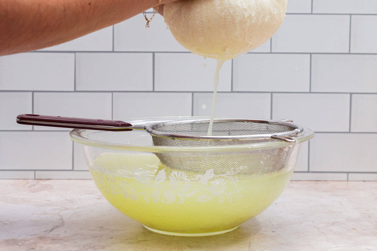 The cheesecloth filled with curds being squeezed over a sieve on a large bowl.