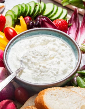 A close up of a bowl of farmer's cheese with a spoon on a platter surrounded by slices of bread and an assortment of vegetables.