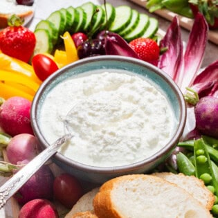 A close up of a bowl of farmer's cheese with a spoon on a platter surrounded by slices of bread and an assortment of vegetables.