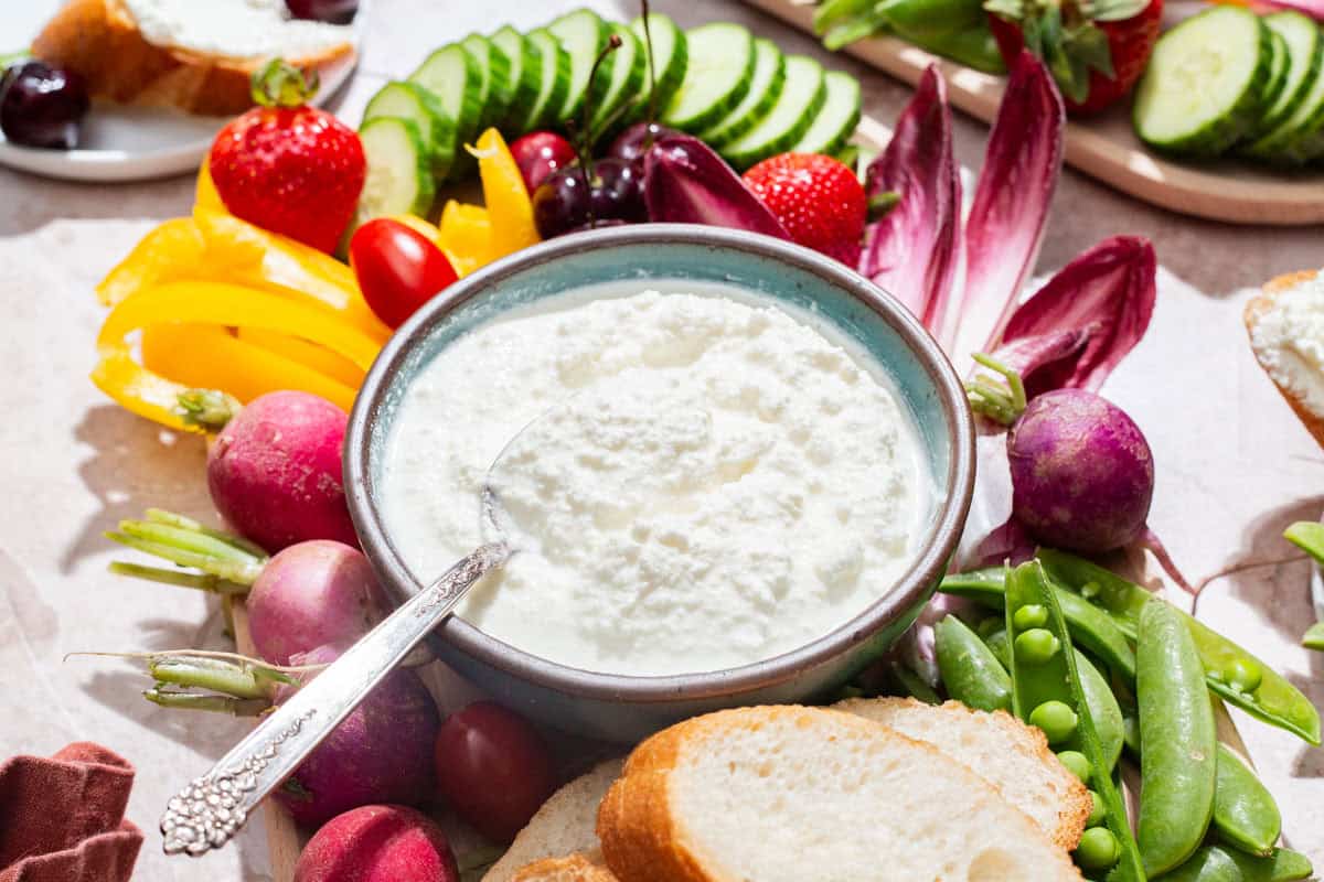 A close up of a bowl of farmer's cheese with a spoon on a platter surrounded by slices of bread and an assortment of vegetables.