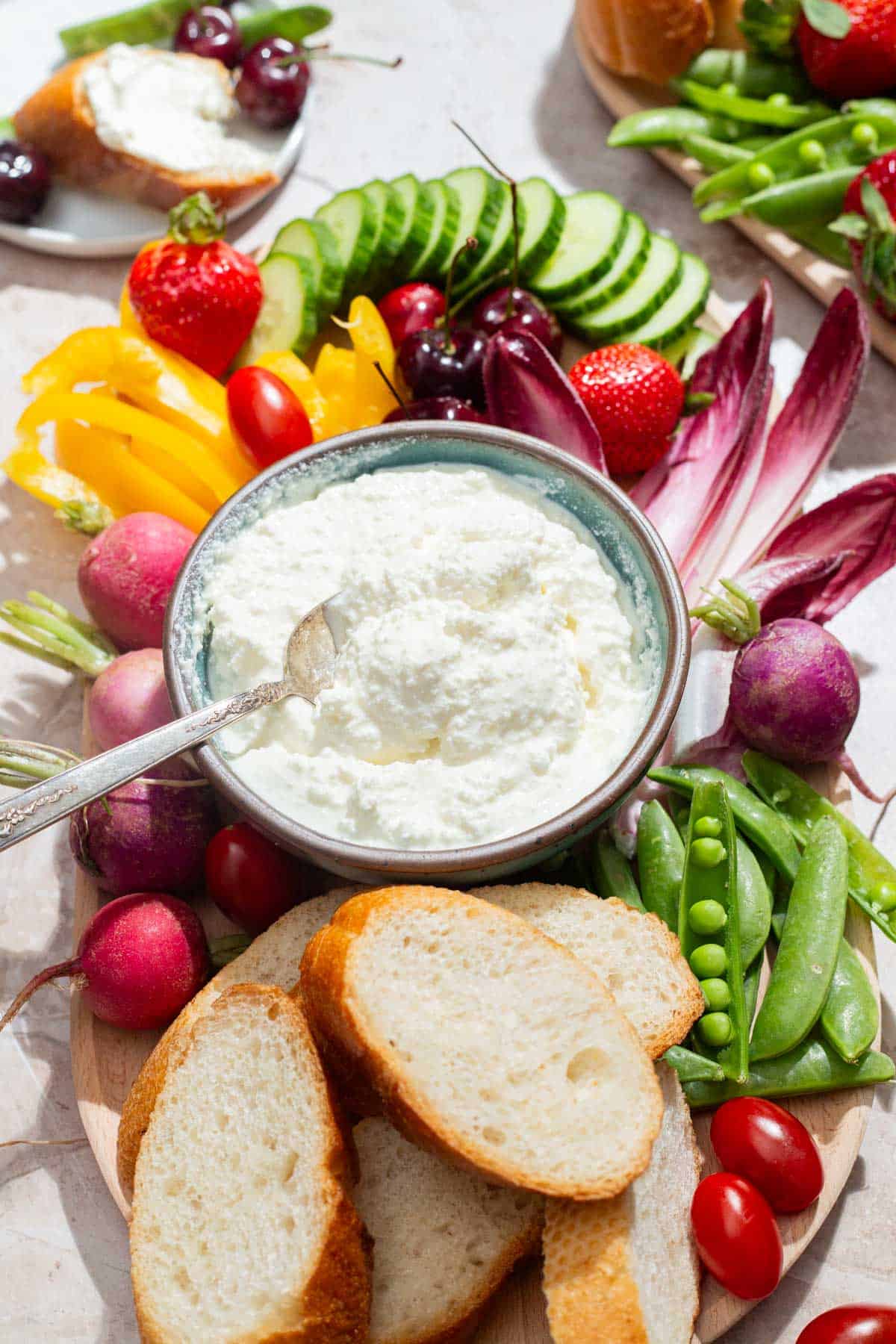 A close up of a bowl of farmer's cheese with a spoon on a platter surrounded by slices of bread and an assortment of vegetables.