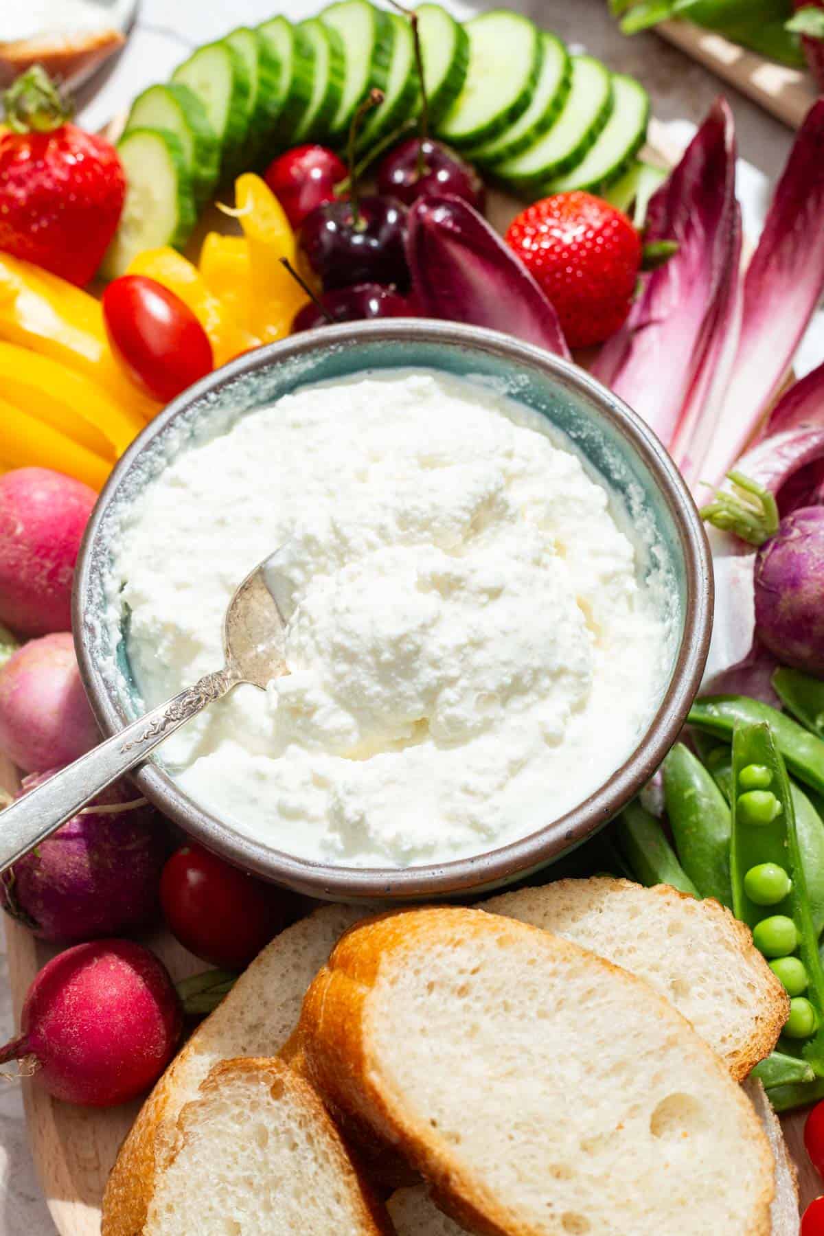 A close up of a bowl of farmer's cheese with a spoon on a platter surrounded by slices of bread and an assortment of vegetables.