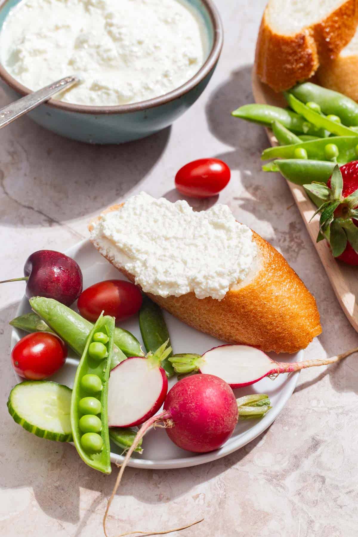 A close up of of farmer's cheese spread on a piece of bread on a plate with an assortment of vegetables. Next to this is the bowl of farmer's cheese with a spoon and a platter with more bread and veggies.