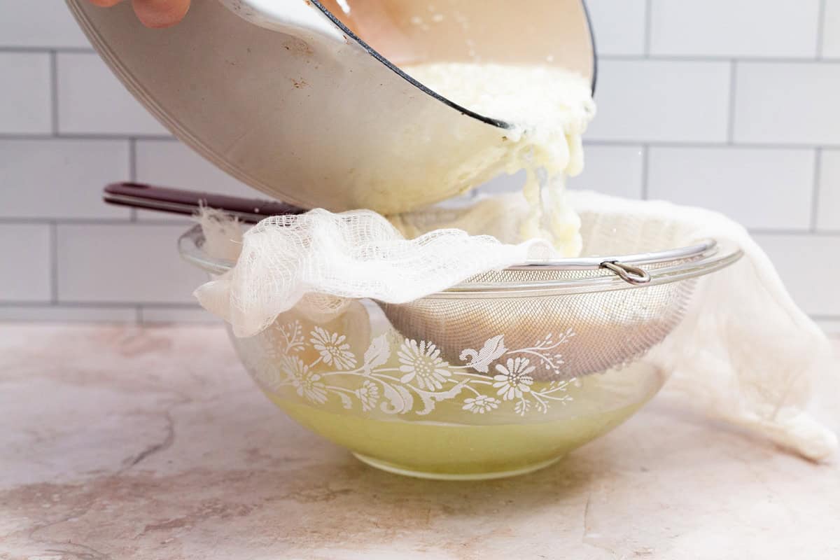 Milk and curds being poured from a pot into a cheesecloth lined sieve over a large bowl.