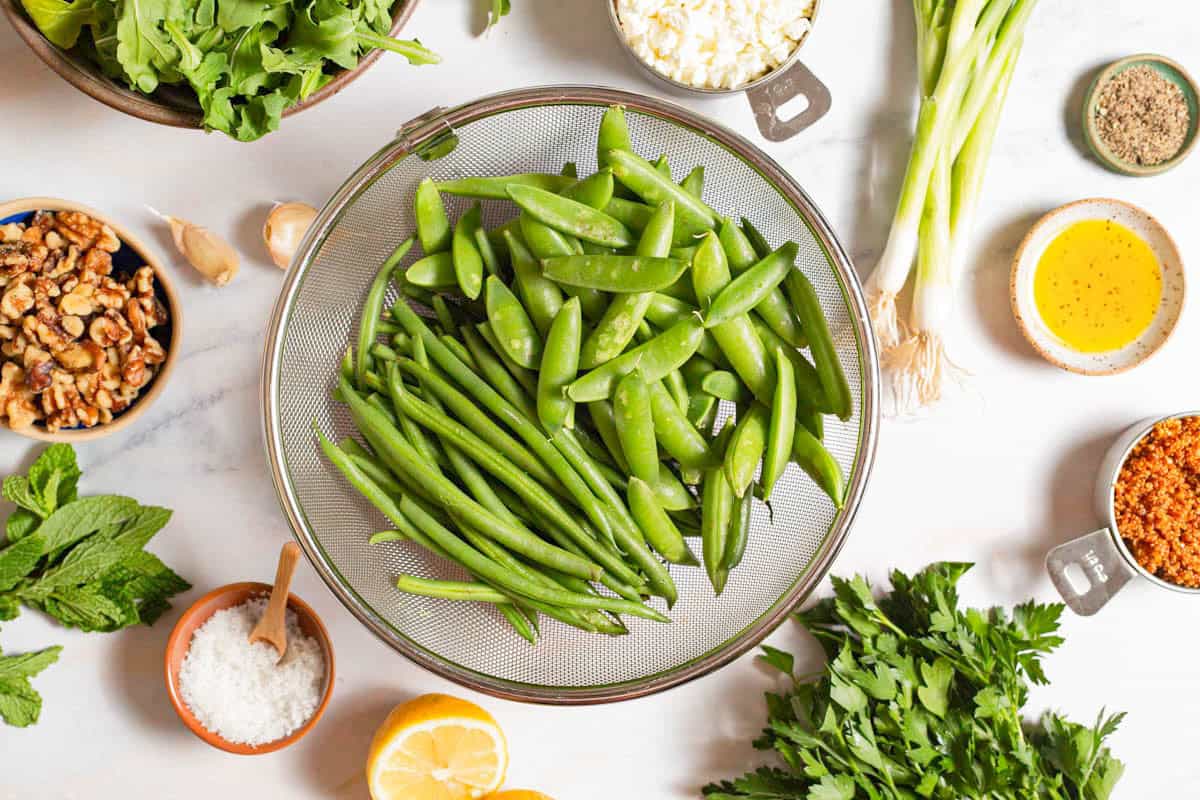 Ingredients for green bean and snap pea salad with crispy quinoa including green beans, snap peas, lemon, garlic, salt, pepper, olive oil, feta cheese, walnuts, scallions, mint, arugula, and crispy quinao.