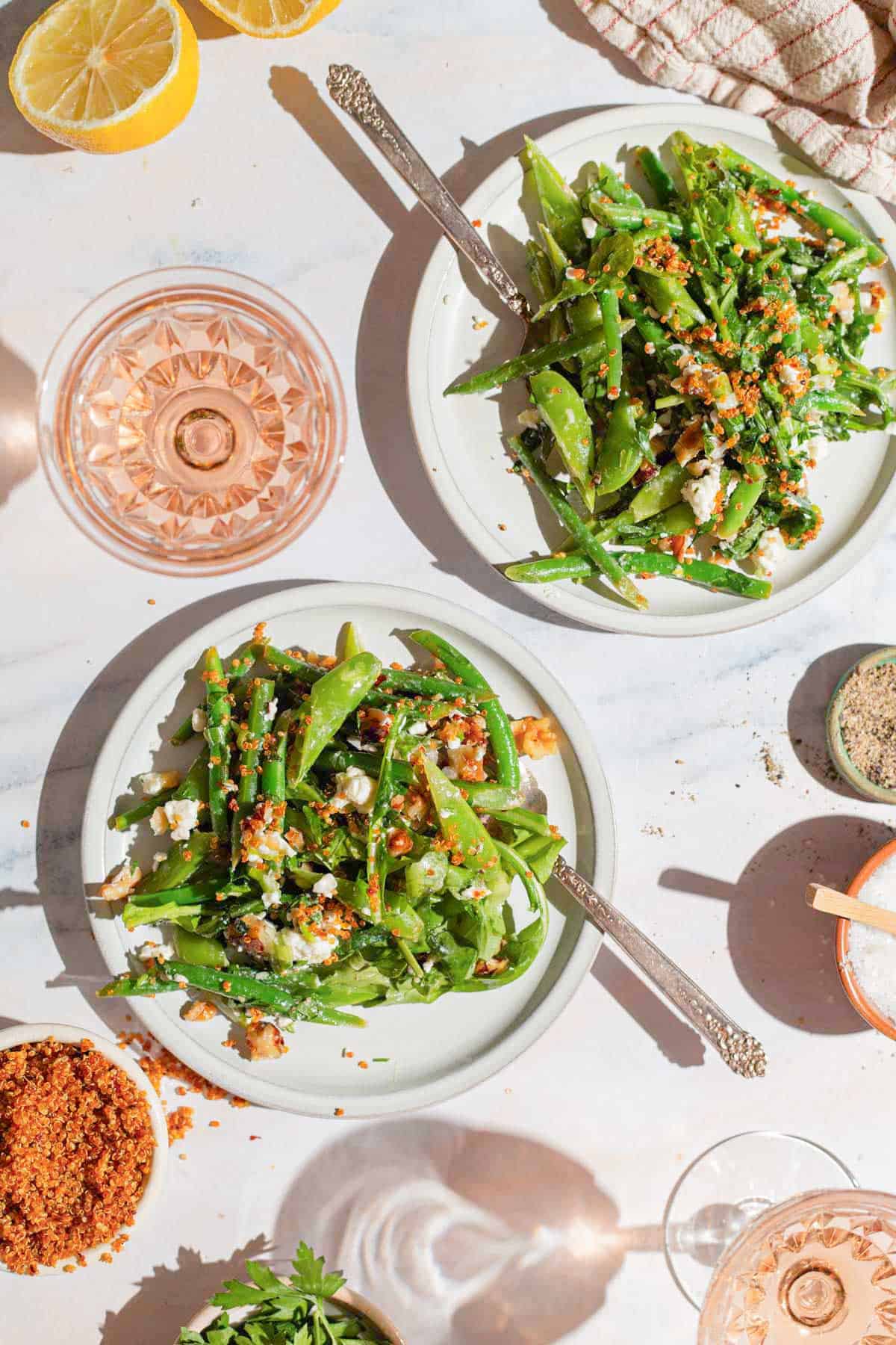 An overhead photo of 2 servings of green bean and snap pea salad with crispy quinoa on plates with forks. Next to these is a kitchen towel, 2 lemon halves, 2 glasses of wine and bowls of quinoa, parsley, salt and pepper.