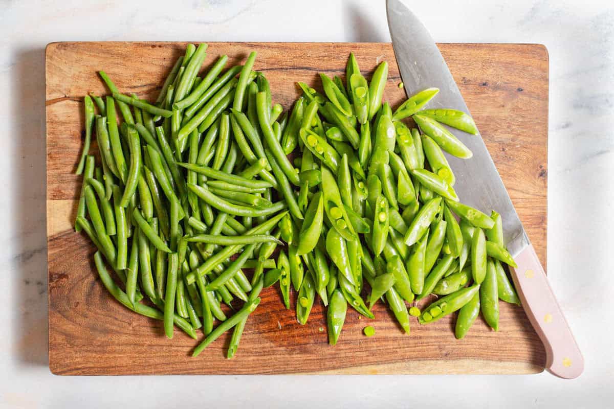 Green beans and snap peas on a cutting board with a knife.