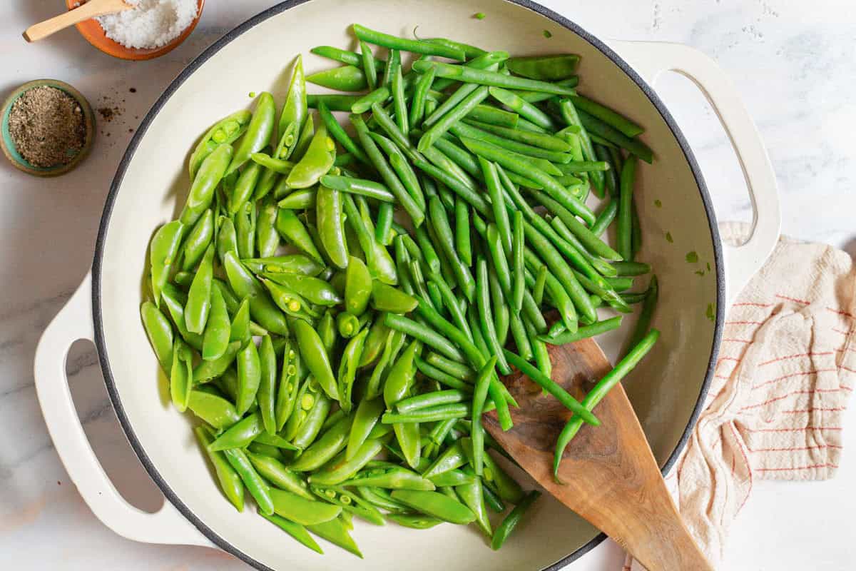 Snap peas and green beans being sauteed in a skillet with a wooden spatula. Next to this is a kitchen towel, and bowls of salt and pepper.