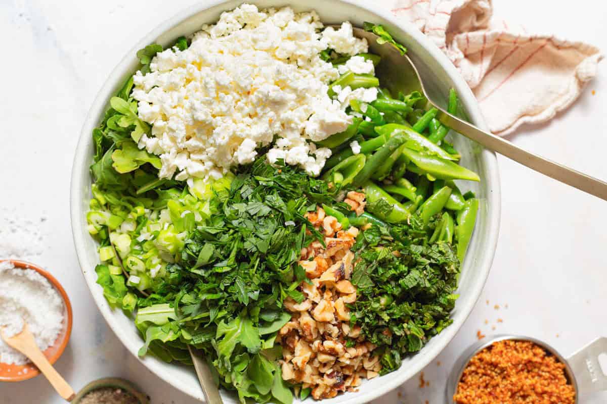 A close up of the ingredients for green bean and snap pea salad in a large bowl with serving utensils before being mixed together. Next to this is a kitchen towel and bowls of salt and crispy quinoa.