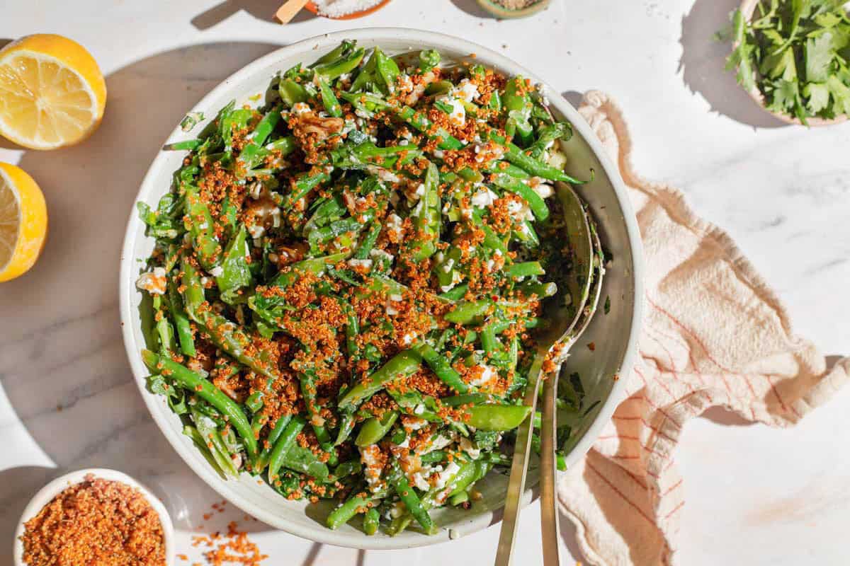 An overhead photo of green bean and snap pea salad with crispy quinoa in a bowl with serving utensils. Next to this is a kitchen towel, two lemon halves, and bowls of parsley and crispy quinoa.