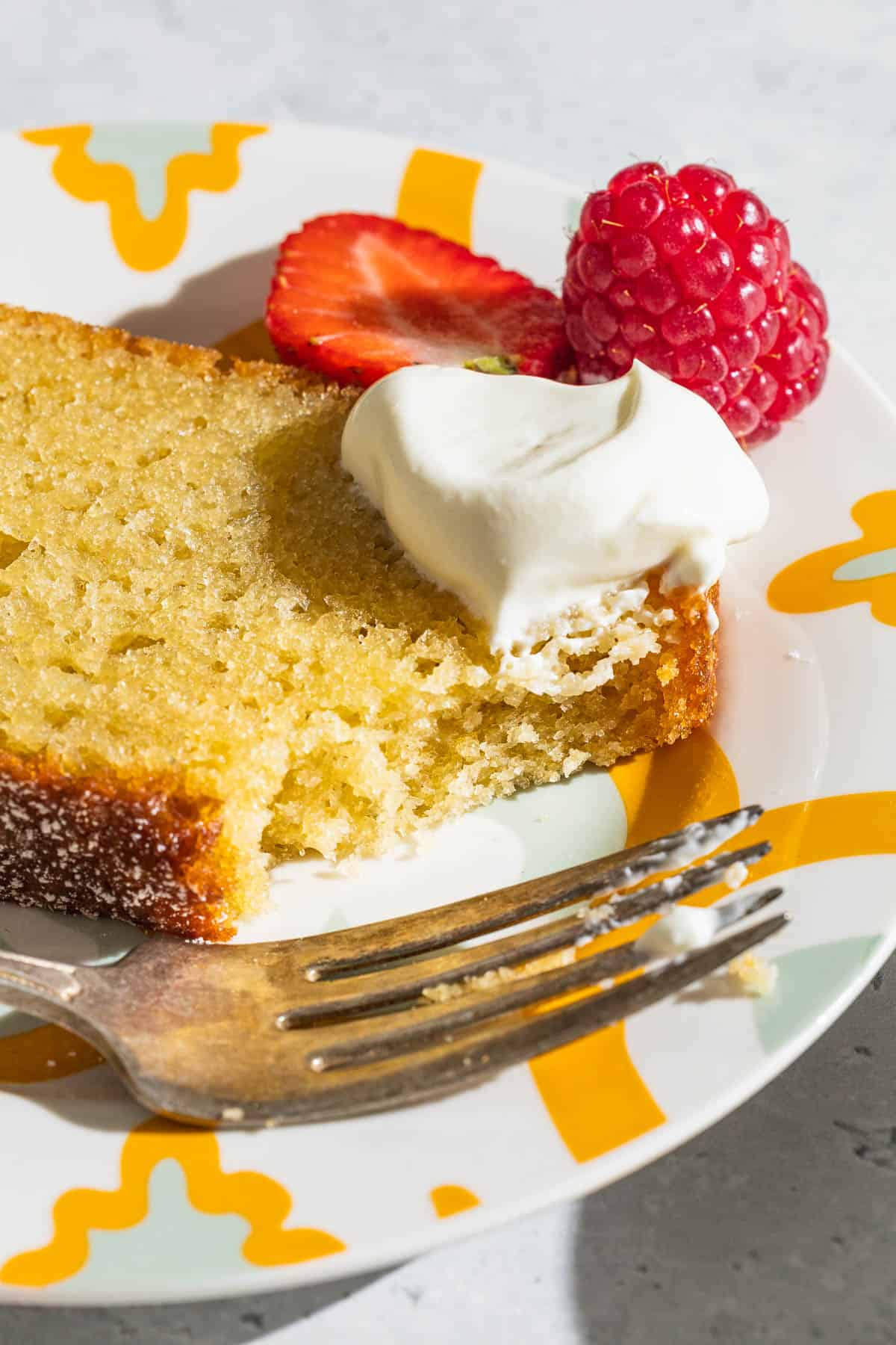 A close up of a slice of yogurt cake on a plate with a dollop of greek yogurt, a strawberry, a raspberry and a fork.