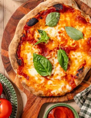An overhead photo of a neapolitan pizza topped with basil leaves on a wooden platter. Next to this is a plate of tomatoes, a kitchen towel, and a bowl of pizza sauce.