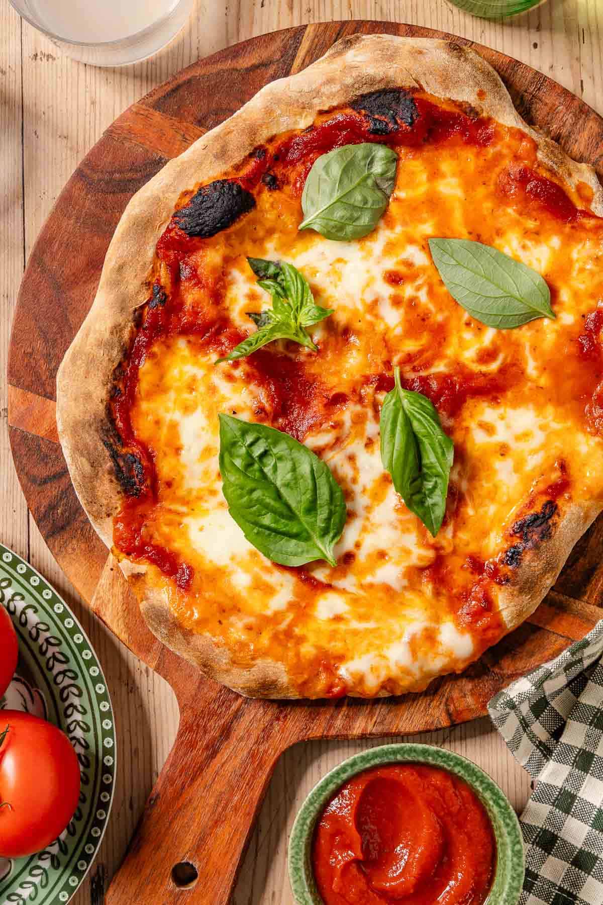 An overhead photo of a neapolitan pizza topped with basil leaves on a wooden platter. Next to this is a plate of tomatoes, a kitchen towel, and a bowl of pizza sauce.