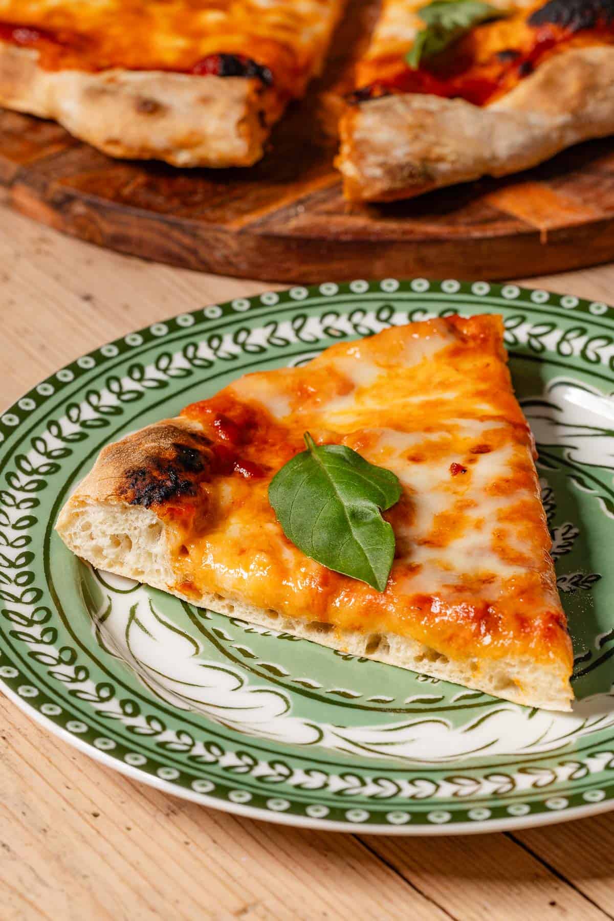 A close up of a slice of neapolitan pizza on a plate with the rest of the pizza on a wooden platter in the background.