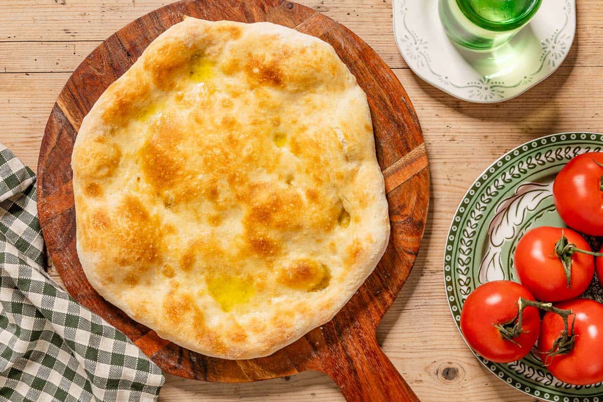 An overhead photo of baked neapolitan pizza dough on a wooden platter. Next to this is a plate with tomatoes, a glass of water on a plate and a kitchen towel.