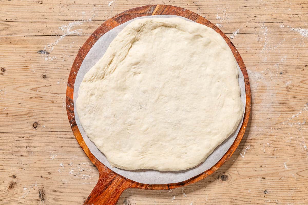 Unbaked neapolitan pizza dough spread on a parchment lined wooden platter.