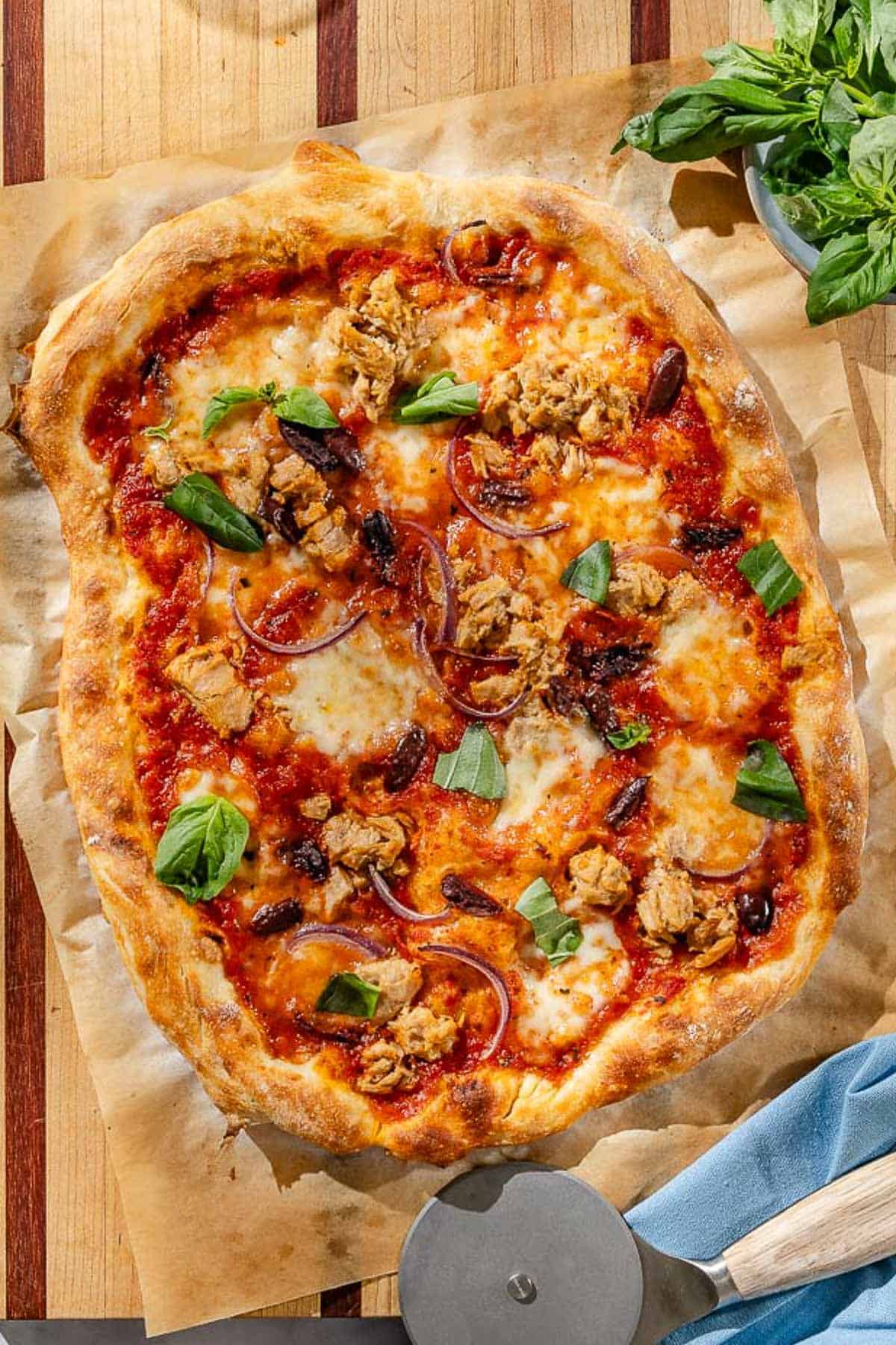 An overhead photo of tuna pizza on a piece of parchment paper on a wooden cutting board. Next to this is a pizza slicer, a cloth napkin, and a bowl of basil.