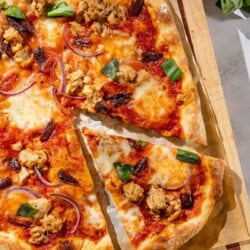 An overhead photo of tuna pizza on a piece of parchment paper on a wooden cutting board, with one slice cut. Next to this is a cloth napkin and a bowl of basil.