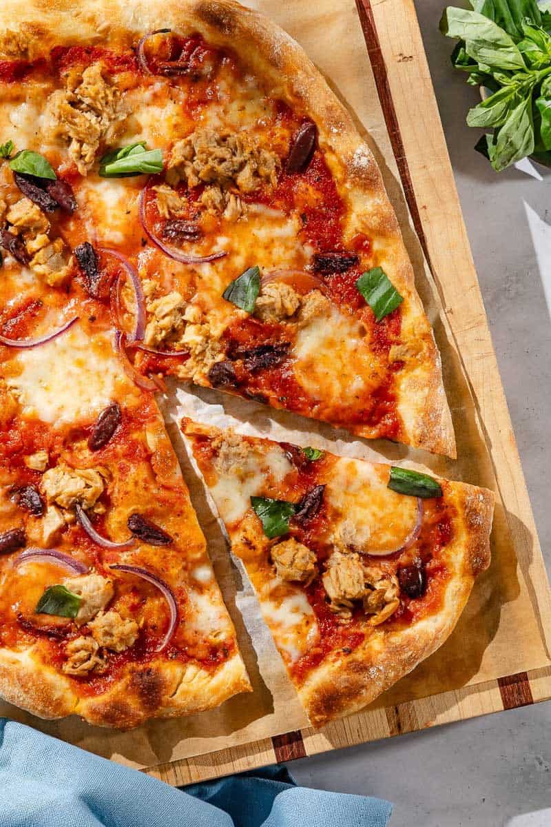 An overhead photo of tuna pizza on a piece of parchment paper on a wooden cutting board, with one slice cut. Next to this is a cloth napkin and a bowl of basil.