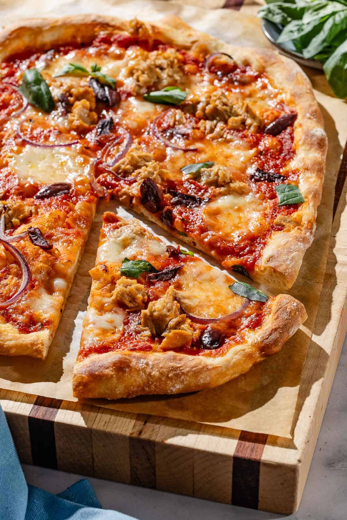 A close up photo of tuna pizza on a piece of parchment paper on a wooden cutting board, with one slice cut. Next to this is a cloth napkin and a bowl of basil.