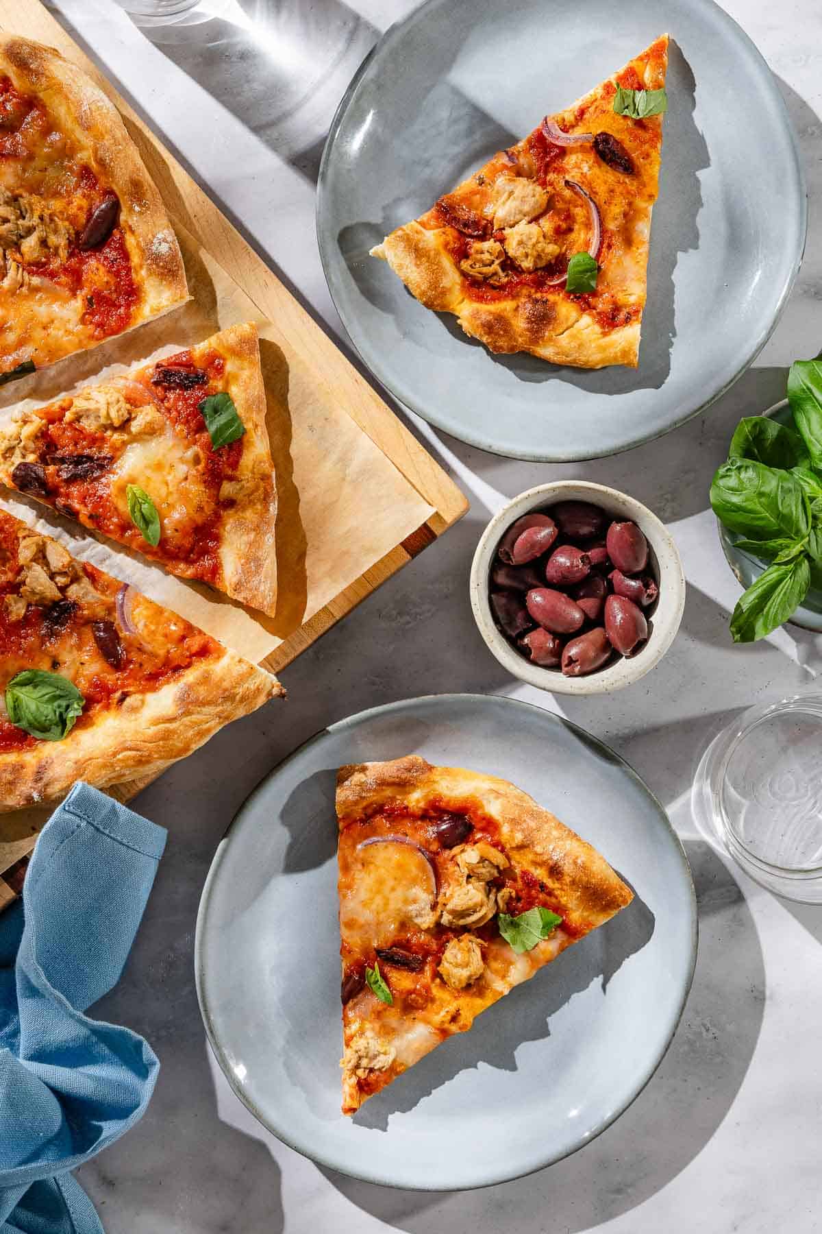 An overhead photo of 2 slices of tuna pizza on 2 plates. Next to this is the rest of the pizza on a wooden cutting board, a cloth napkin, bowls of black olives and basil and a glass of water.