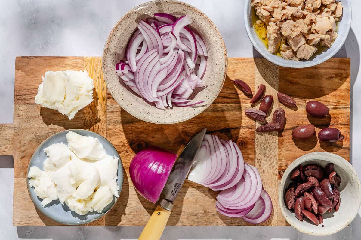 A cutting board with a knife and the topping for tuna pizza including mozzarella cheese, red onion, black olives and tuna.