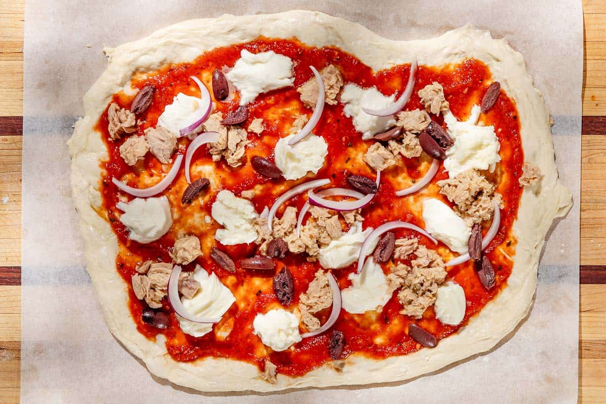 A close up photo of an unbaked tuna pizza on parchment on a wooden cutting board.