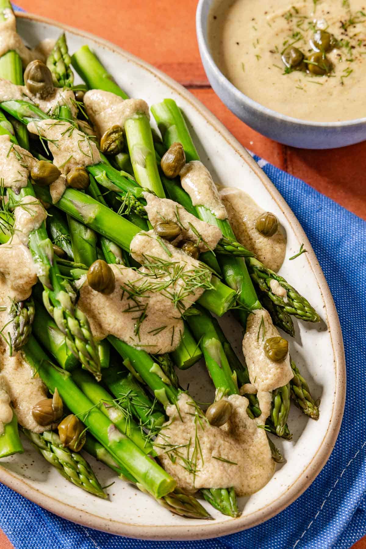 A close up of asparagus topped with tonnato sauce, dill and capers on a serving plater. Next to this is a bowl of the tonnato sauce.