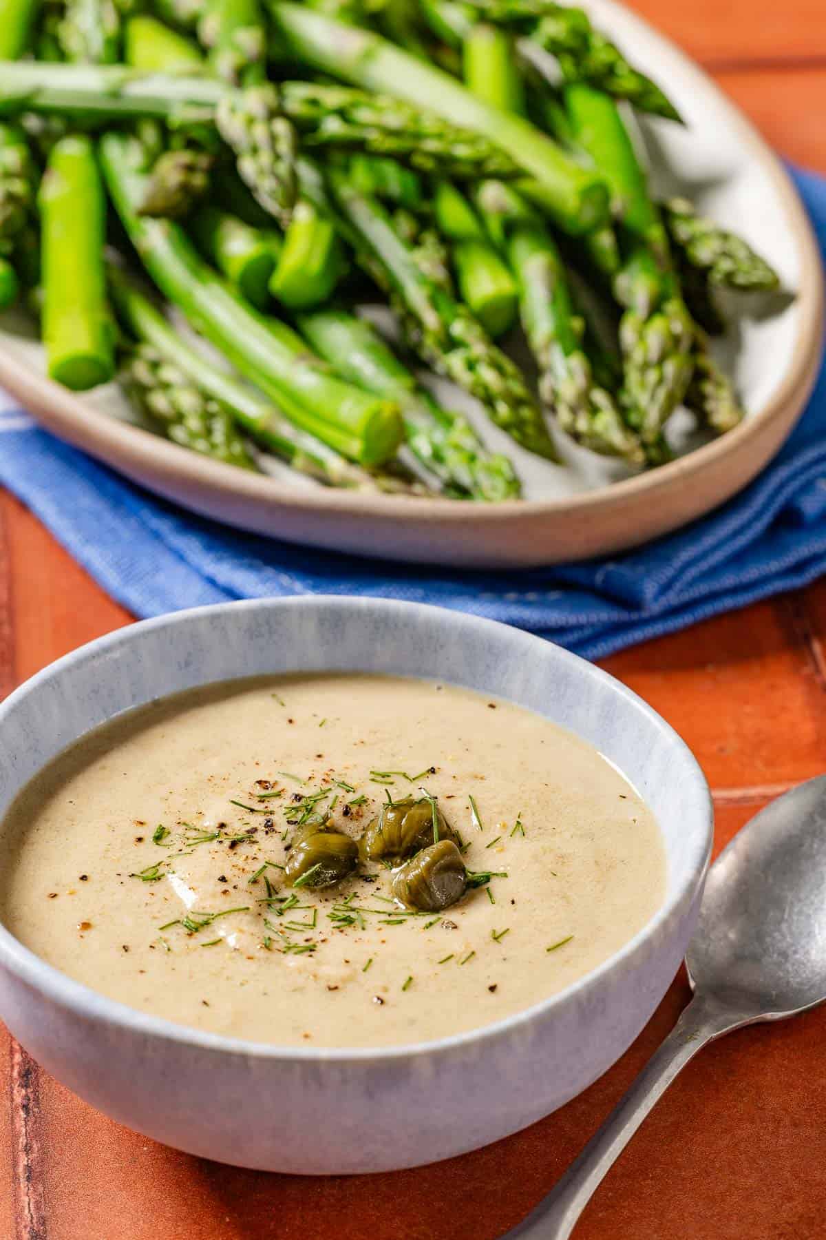 A close up of asparagus topped with tonnato sauce, dill and capers on a serving plater. Next to this is a bowl of the tonnato sauce and a spoon.