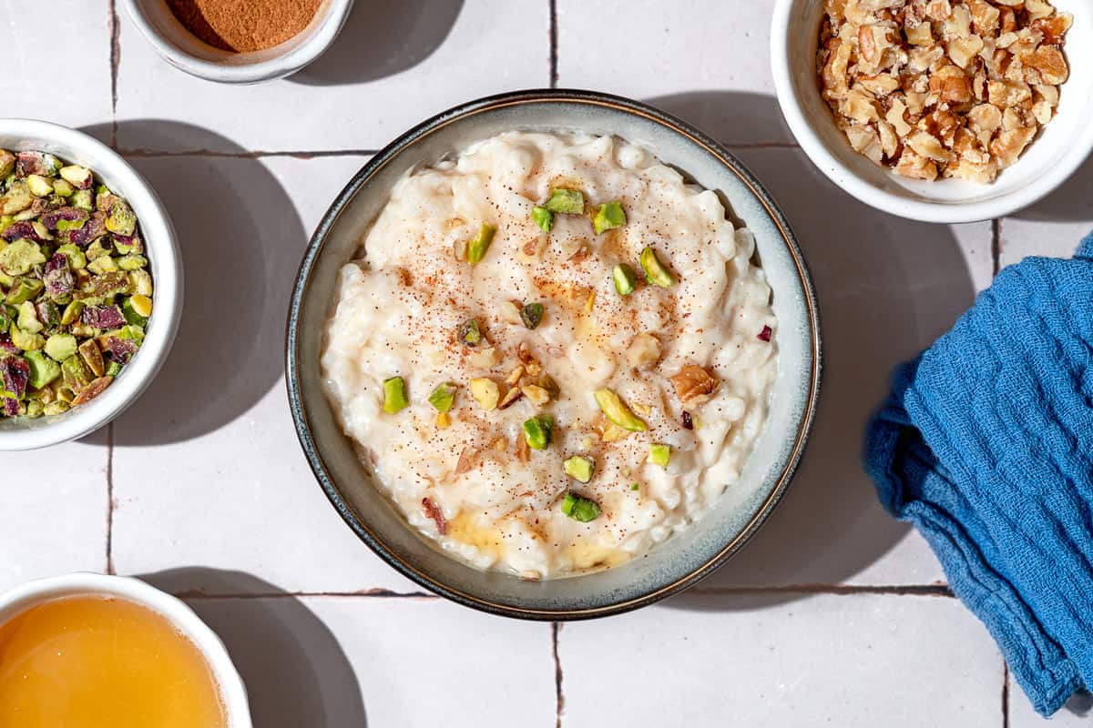 An overhead photo of easy rice pudding garnished with pistachios, walnuts, cinnamon and a drizzle of honey. Next to this is a kitchen towel and bowls of cinnamon, crushed pistachios, crushed walnuts and honey.