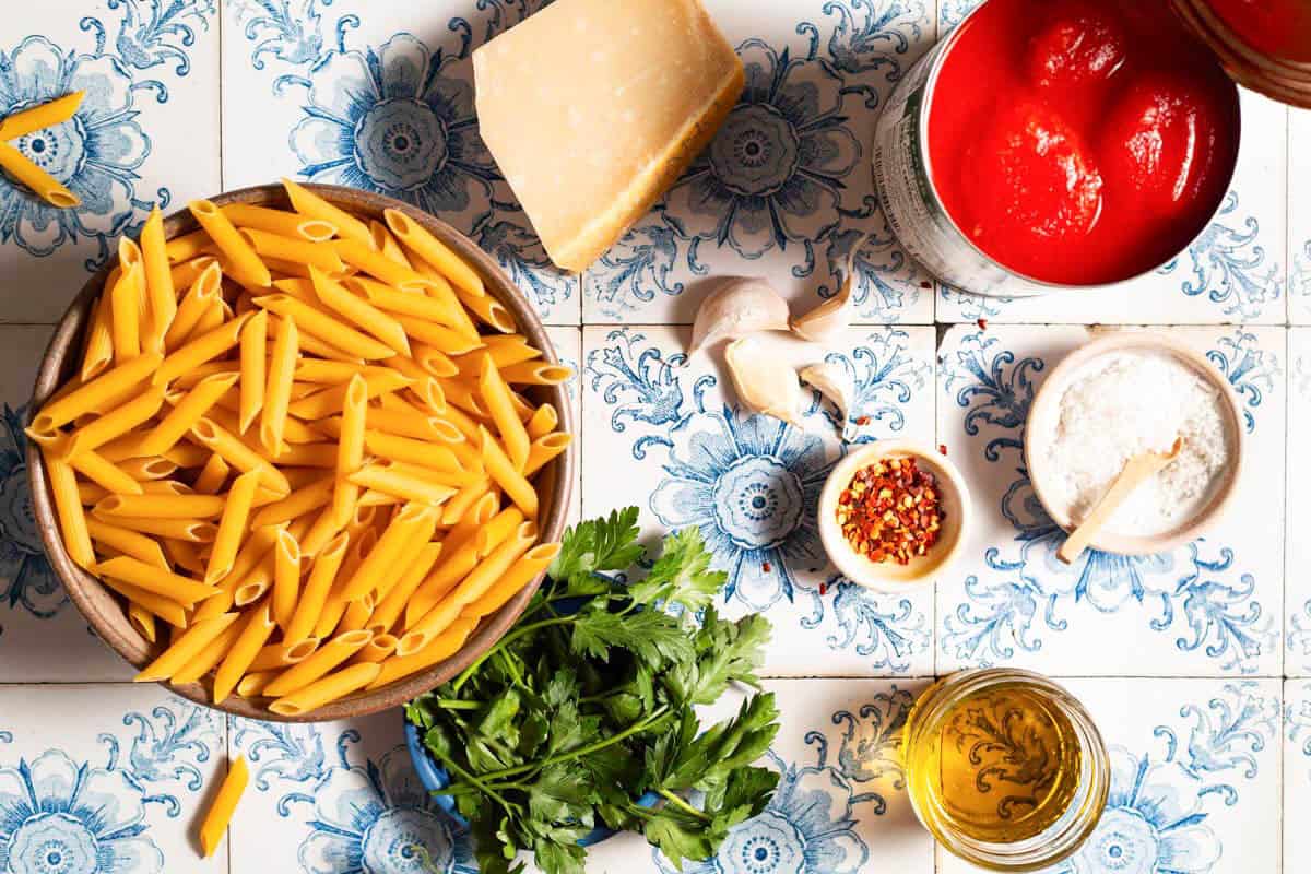 Ingredients for arrabbiata sauce pasta including penne, garlic, olive oil, red pepper flakes, canned whole tomatoes, salt, parsley, and parmesan cheese.