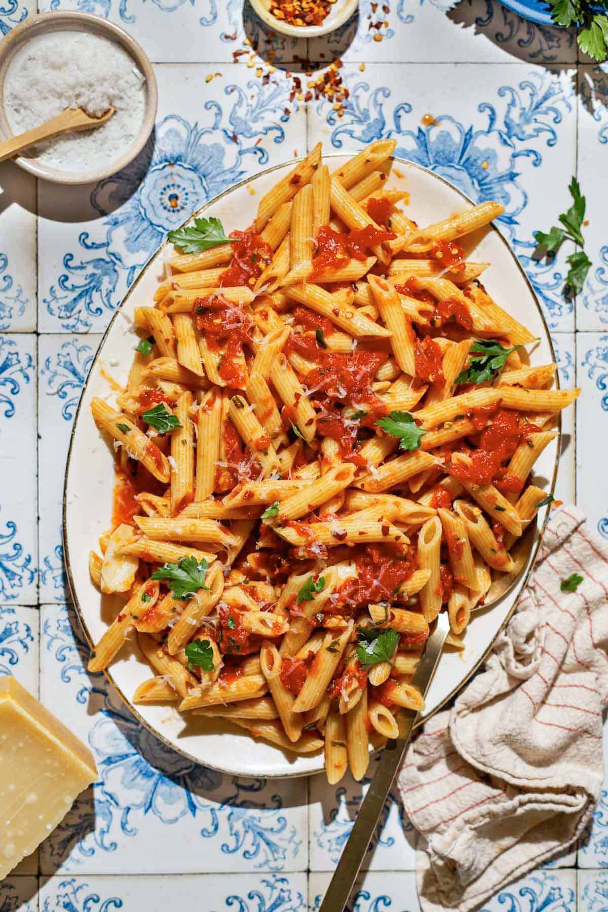 An overhead photo of arrabbiata sauce pasta on a serving platter with a spoon. Next to this is a kitchen towel, a wedge of parmesan cheese, and bowl of salt, red pepper flakes and parsley.