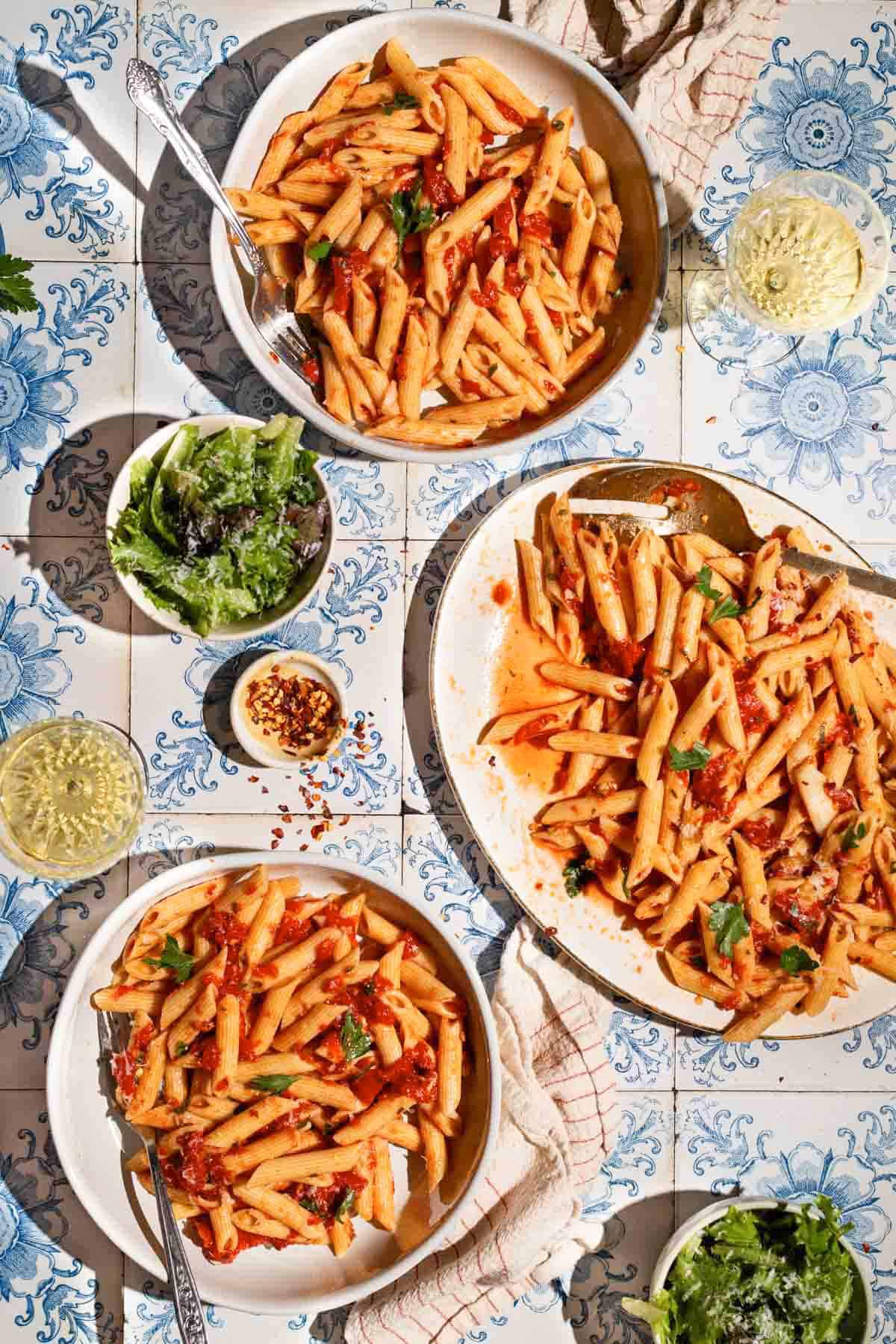 An overhead photo of 2 servings of arrabbiata sauce pasta in bowls with forks, the rest of the pasta on a serving platter with a spoon, 2 glasses of white wine, 2 bowls of salad and a bowl of red pepper flakes.