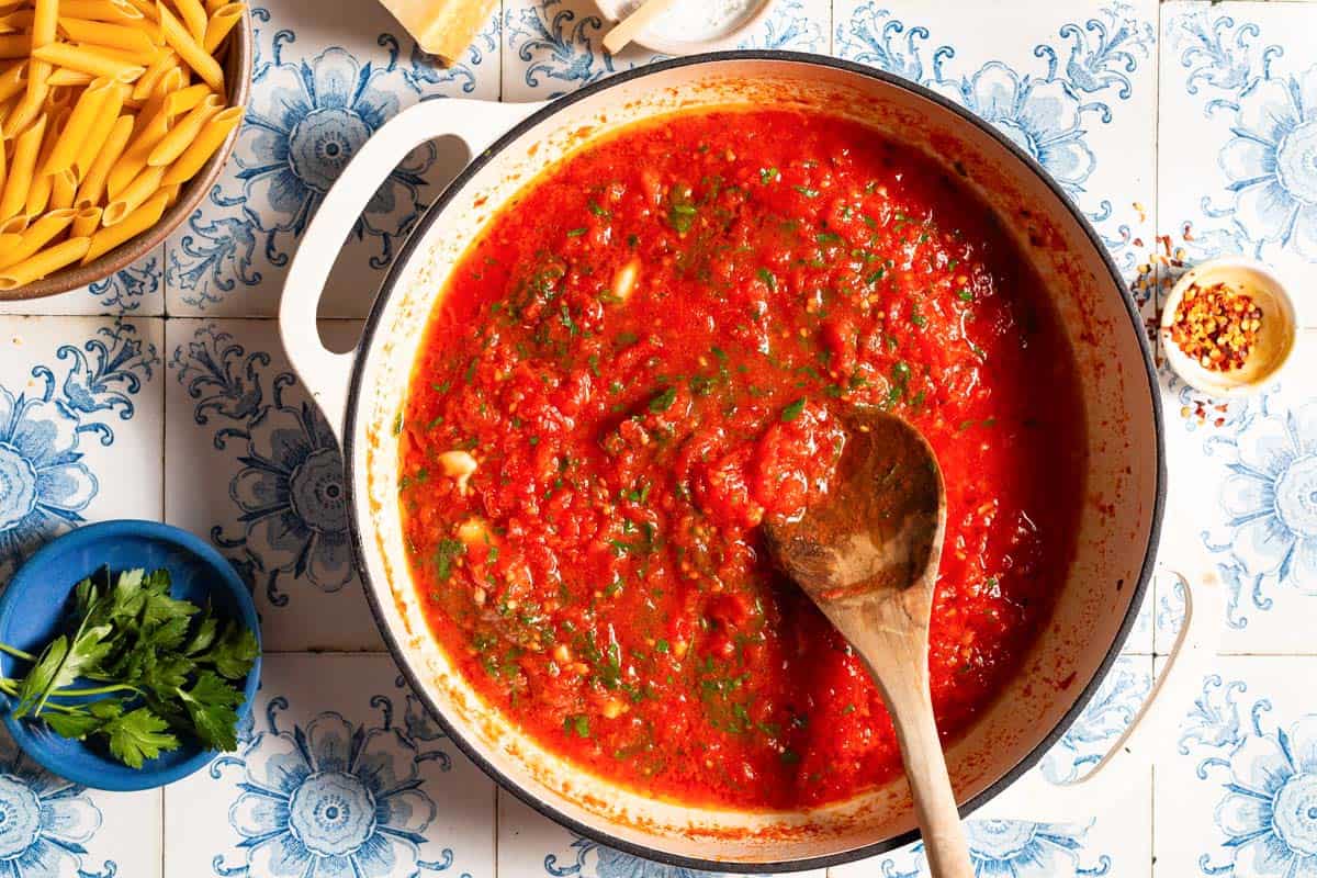 Arrabbiata sauce cooking in a skillet with a wooden spoon. Nest to this are bowls of parsley, pasta, salt and red pepper flakes.